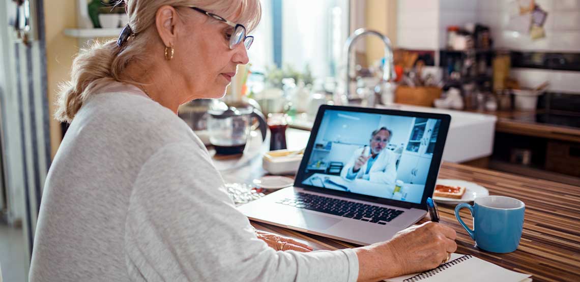 A woman sitting in front of her laptop at home taking notes during a telehealth appointment.