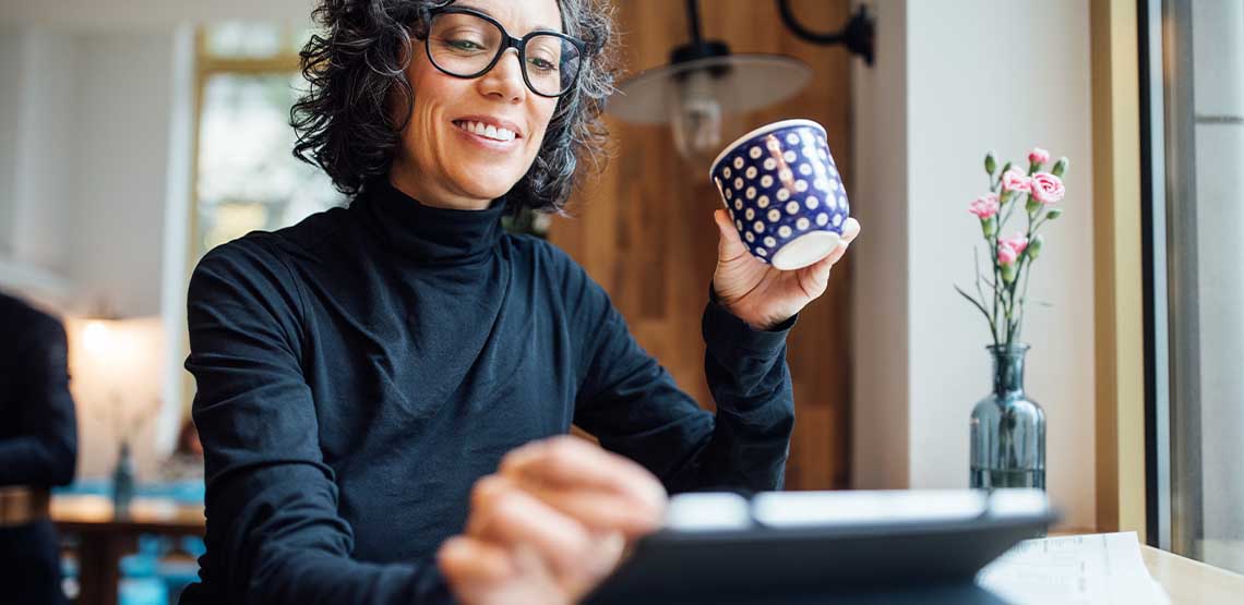 A woman holding a mug and wearing glasses while browsing on her tablet.